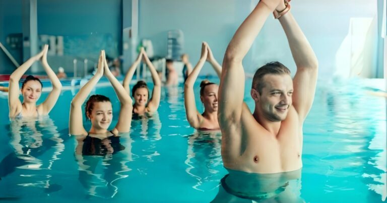 A group of individuals in a swimming pool, engaged in a synchronized exercise or swimming activity. Each person has their arms raised and joined together above their heads, forming a pointed shape. The setting suggests an indoor pool due to the lighting and reflections on the water surface.